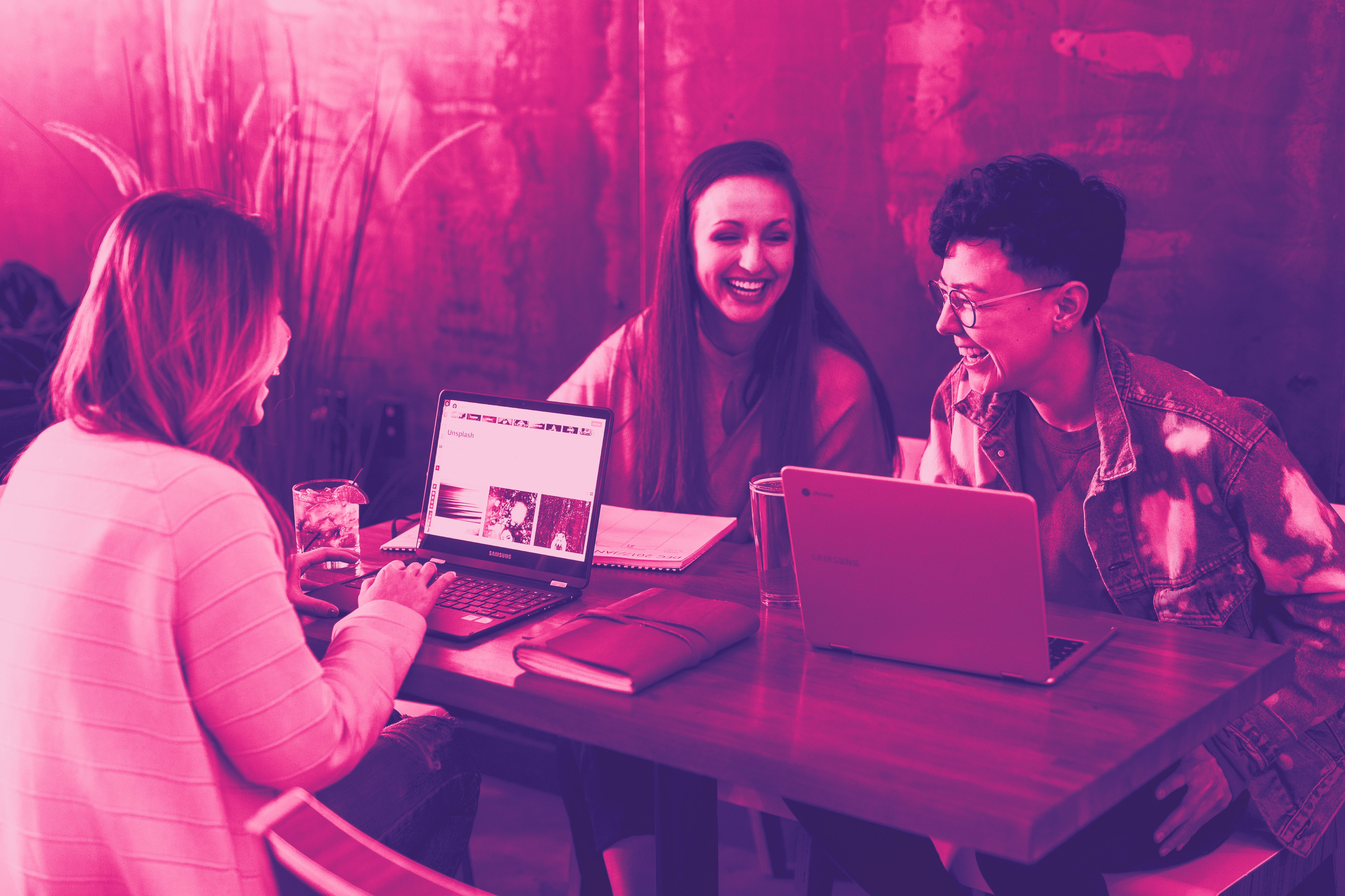 Three women sit at a workstation with computers and laugh, joking with each other. With pink filter