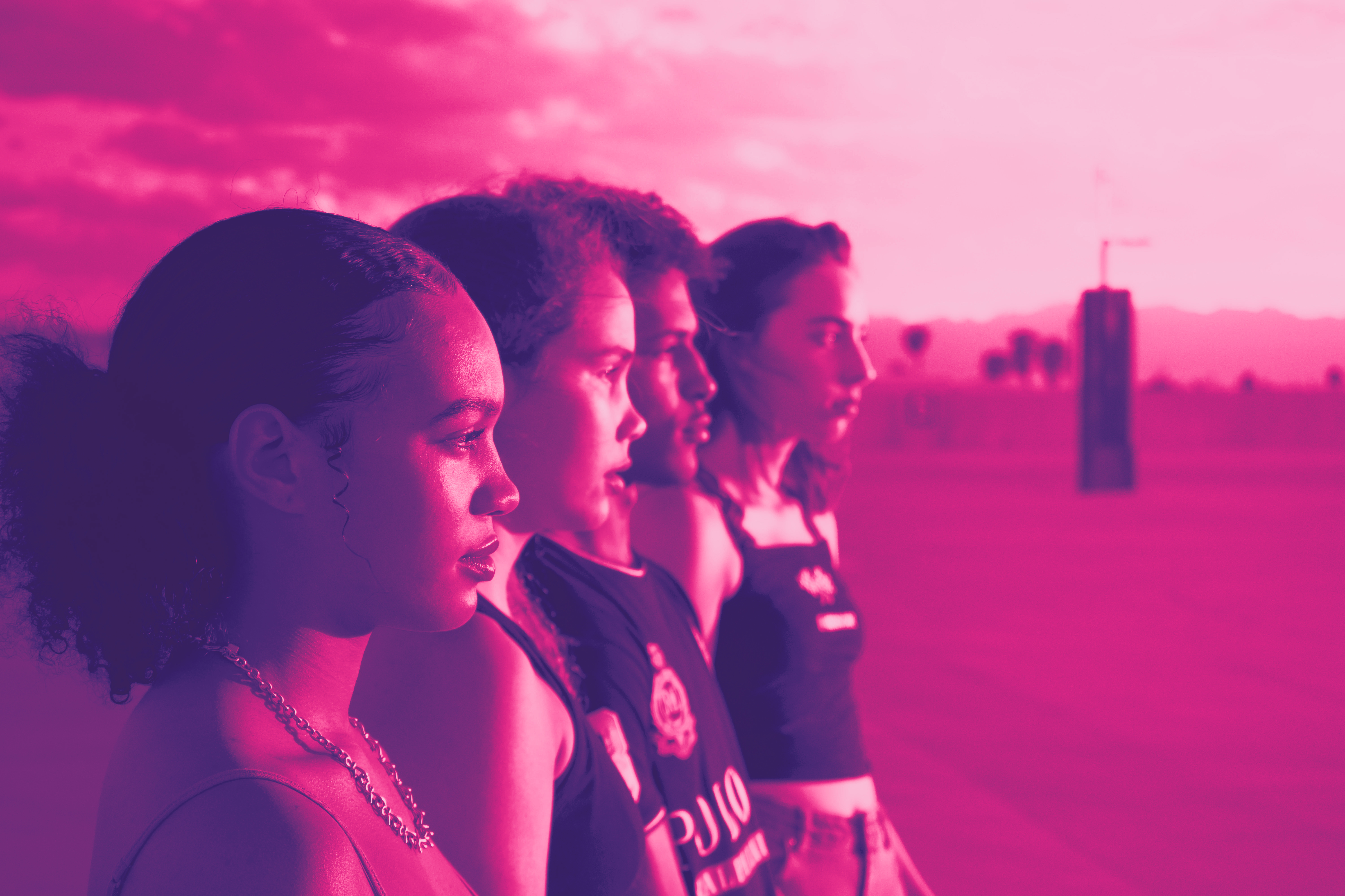 Four young people stand in a row in profile, their eyes fixed on their right. With pink filter