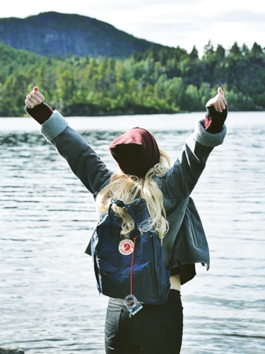 Kvinna framför en stor sjö, med en fjällräven ryggsäck på ryggen, sträcker upp armarna 
