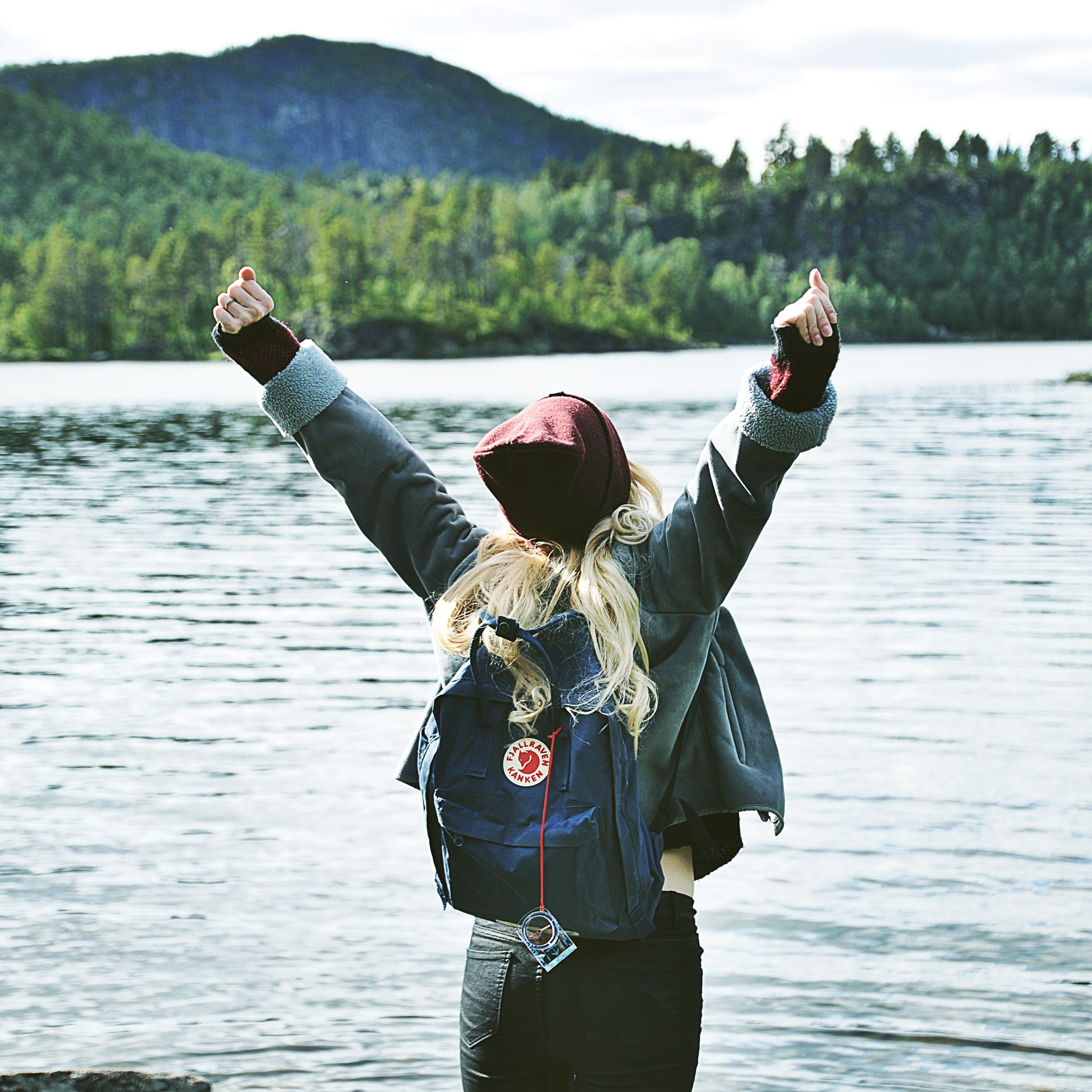 Kvinna framför en stor sjö, med en fjällräven ryggsäck på ryggen, sträcker upp armarna 